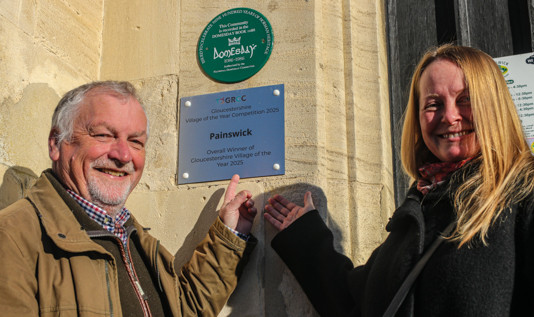 Parish Councillors  Adrian Gyde and Fiona Mackenzie-Jenkins point to the Gloucestershire Village of the Year Winner's plaque on the wall of the Parish Hall 